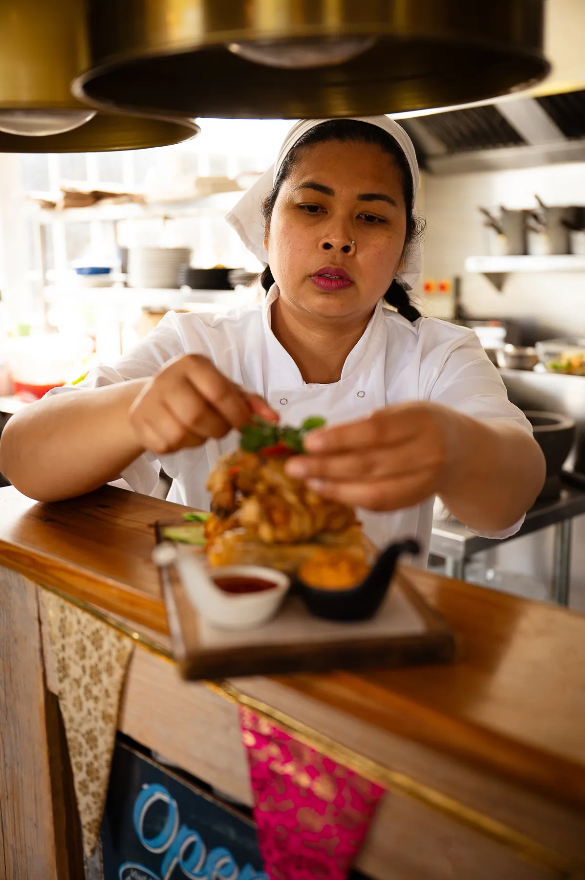 A chef garnishing a finished plate under the heat lamp at the pass