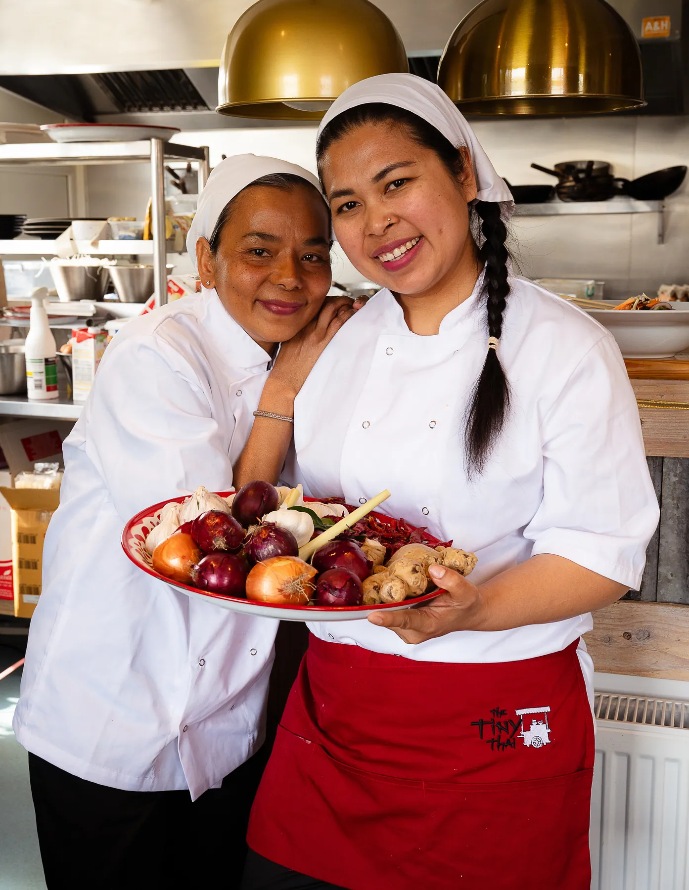 Two chefs in whites presenting a finished dish together in The Tiny Thai kitchen