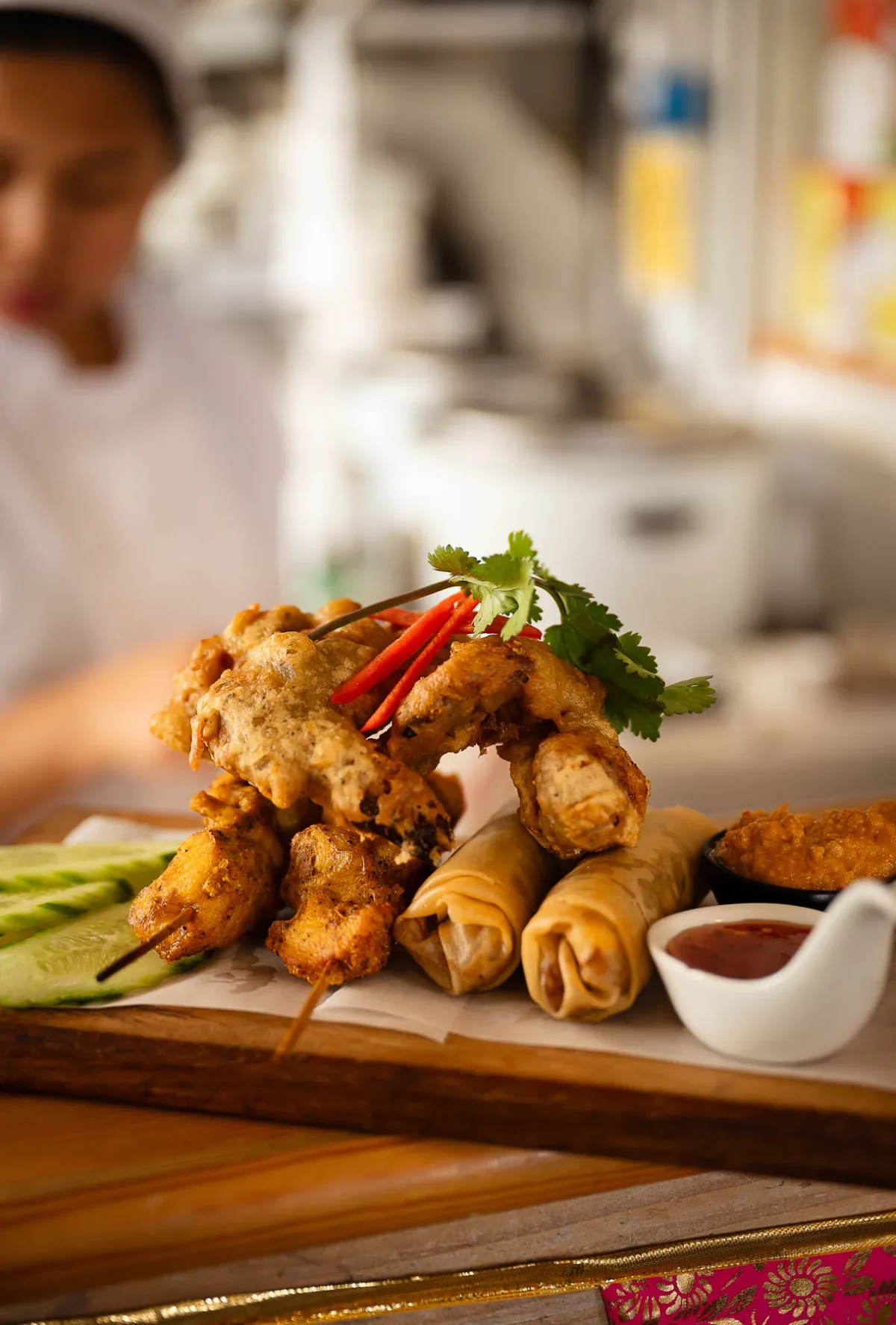 A wooden board of spring rolls, chicken satay skewers and wings with dipping sauces