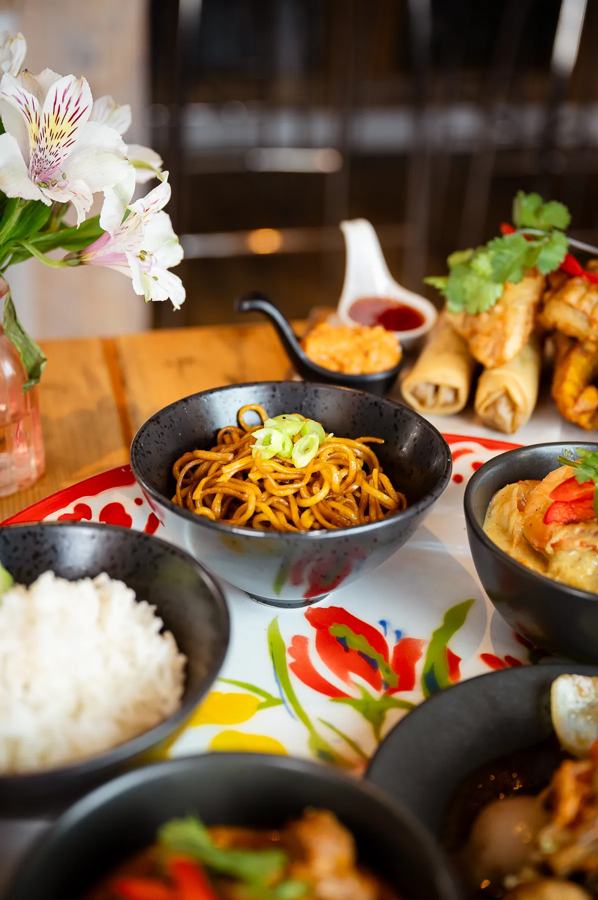 A long table laid with a shared spread of Thai dishes beside a bright window