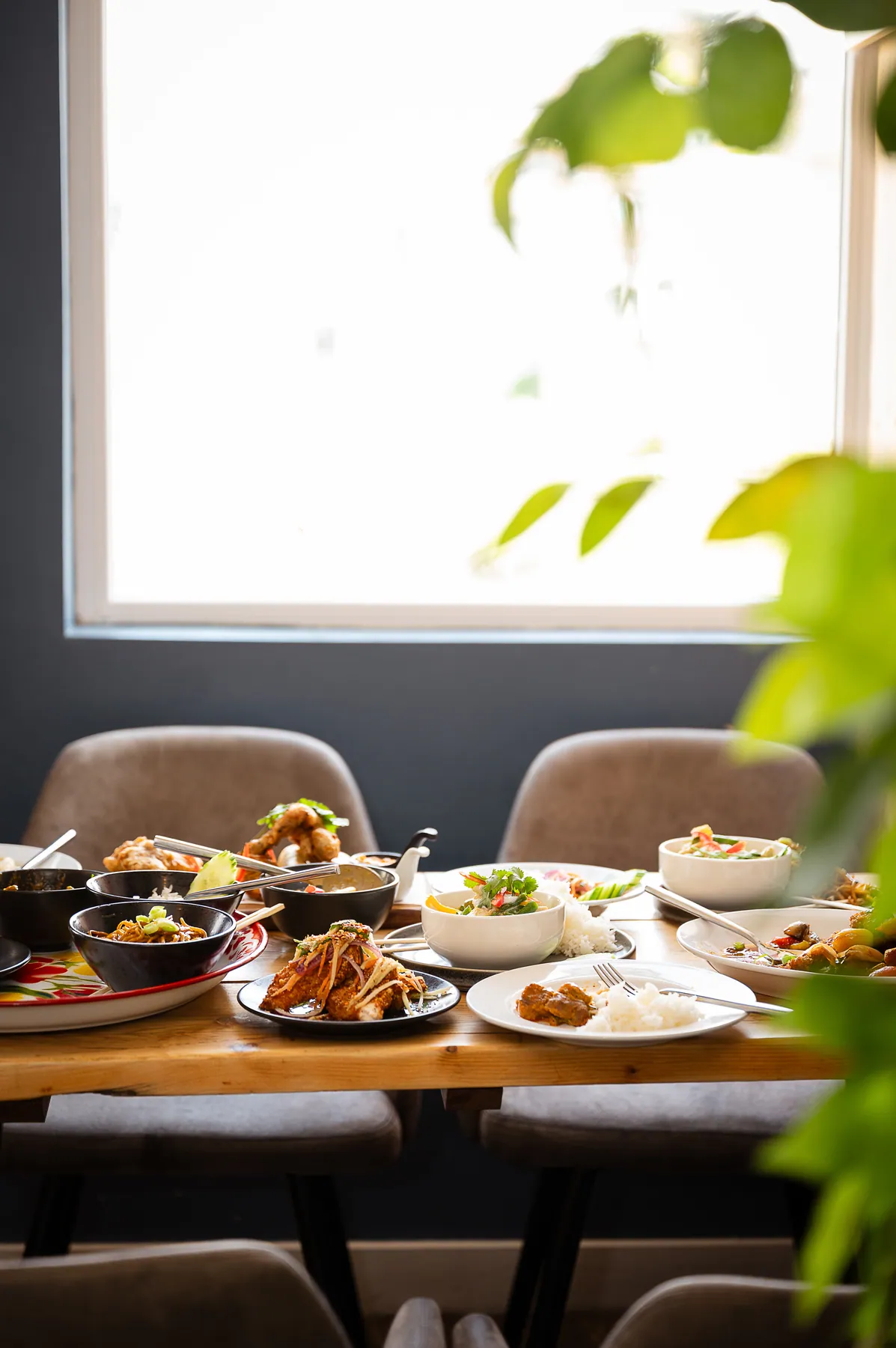 A long table laid with Thai dishes beside a bright window